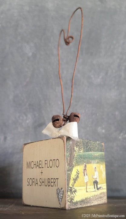 Custom engagement block ornament featuring couple names “Michael Floto + Sofia Shubert” and proposal photo scene by a lake. Accented with glitter heart, ivory ribbon bow, and rusty wire heart hanger.